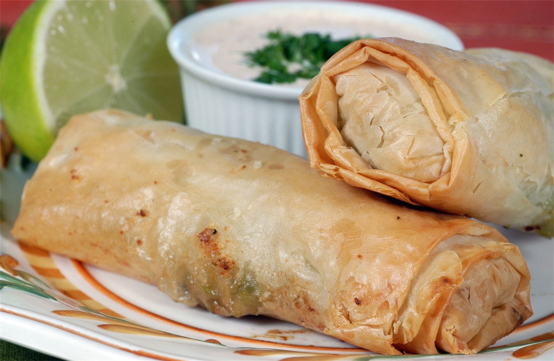 Two crispy spring rolls on a plate, one cut open showing a flaky interior, with lime wedge and dipping sauce. Two crispy spring rolls on a plate, one cut open showing a flaky interior, with lime wedge and dipping sauce.